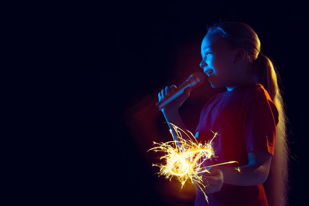 Caucasian girl's portrait on dark studio background in neon light. Beautiful female model with speaker and sparkler. 