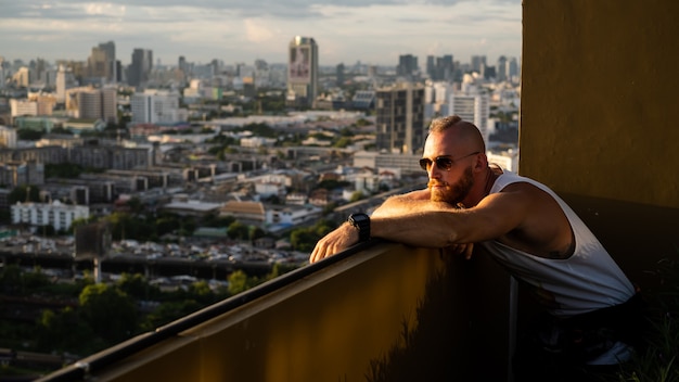 Caucasian handsome bearded brutal man enjoying sunset and view on Bangkok city from high floor