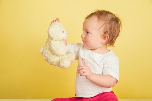 Caucasian little girl, children isolated on yellow studio background. Portrait of cute and adorable kid, baby playing with teddy bear.