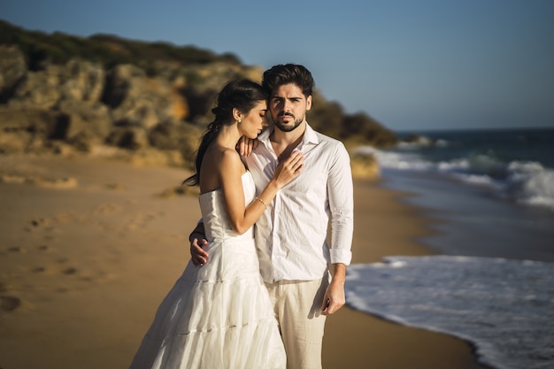 Free photo caucasian loving couple wearing white clothes and hugging in the beach during a wedding photoshoot