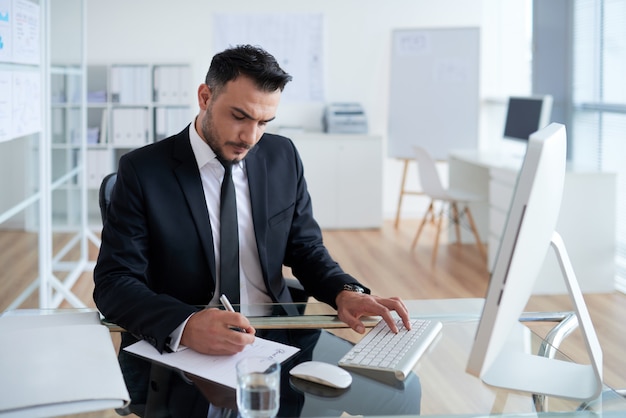 Caucasian man in business suit sitting in office, working on computer and writing on paper