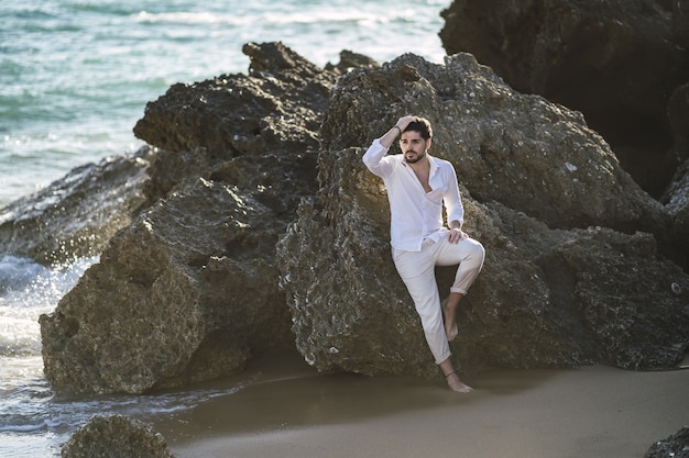 Free photo caucasian man wearing white clothes sitting on the stone on the beach