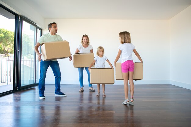 Caucasian parents and two girls holding carton boxes and standing in empty living room