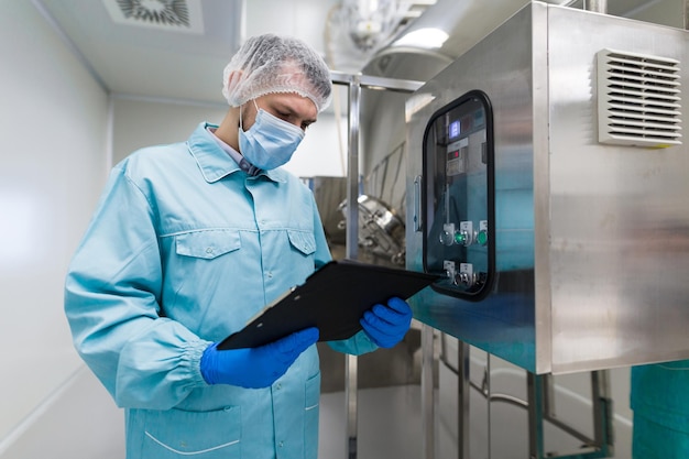 Free photo caucasian scientist in blue lab uniform stand near big steel machine with control panel make notes about machine