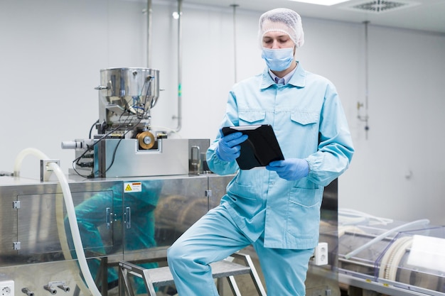 Free photo caucasian scientist man in blue lab suit work with big metal machine in clean room stand on chromed ladder look at camera