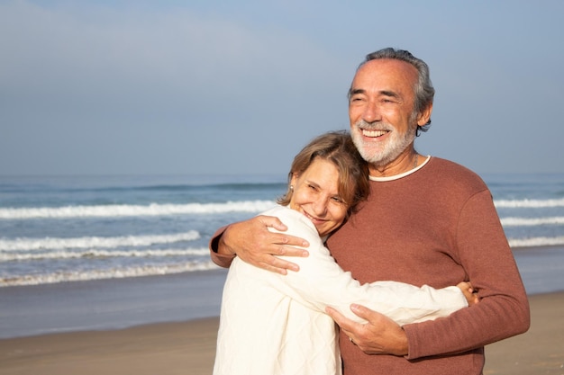 Caucasian senior couple spending time at seashore. Grey-haired man with beard smiling happily while hugging his loving wife. Seascape background. Medium shot. Relationship, travel, retirement concept