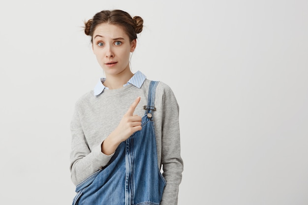 Caucasian woman 20s wondering about what she see showing index finger sideways raising eyebrow. Girl with fashionable odango hairstyle suspiciously showing on white wall . Copy space