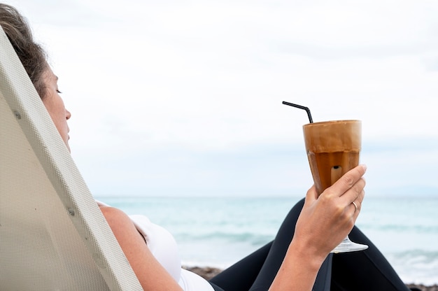 Caucasian woman holding coffee drink on a beach with foam and drinking straw with sea