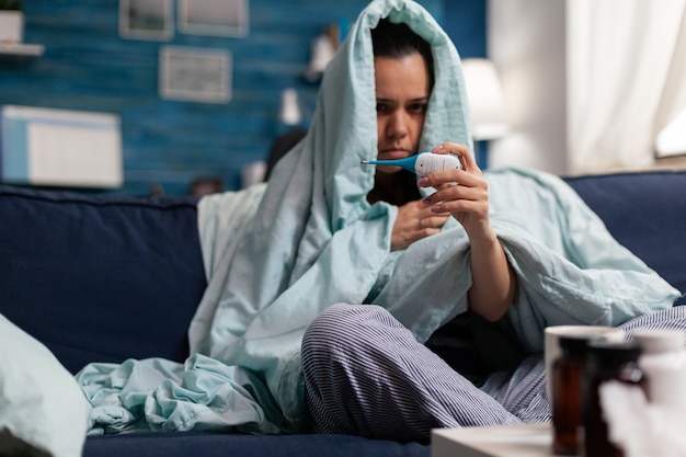 Caucasian woman measuring temperature ill at home with a thermometer. Person feeling sick cold unwell, checking fever and symptoms of disease flu infection. Resting adult with headache