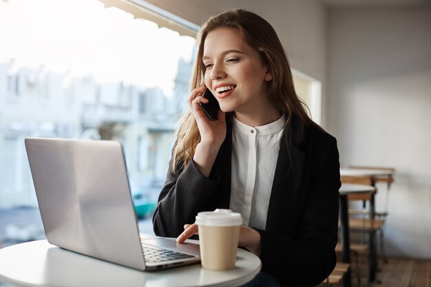 caucasian woman sitting in cafe, drinking coffee, talking on smartphone, looking at laptop screen with broad smile