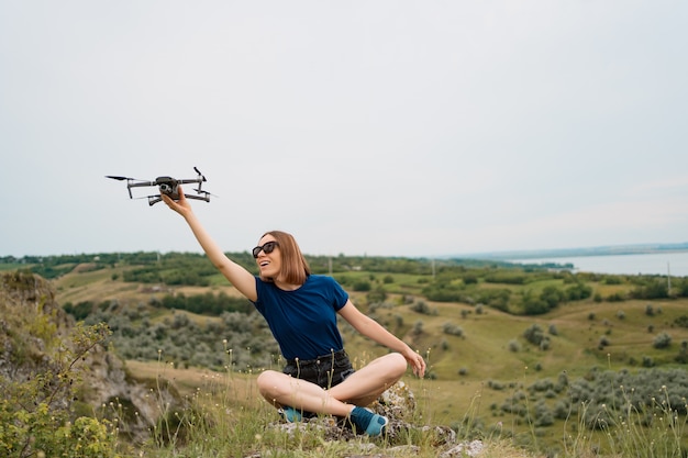 A Caucasian woman with a drone in her hand, sitting on a green rocky hill with sky in background