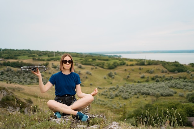 Free photo a caucasian woman with a drone in her hand, sitting on a green rocky hill with sky