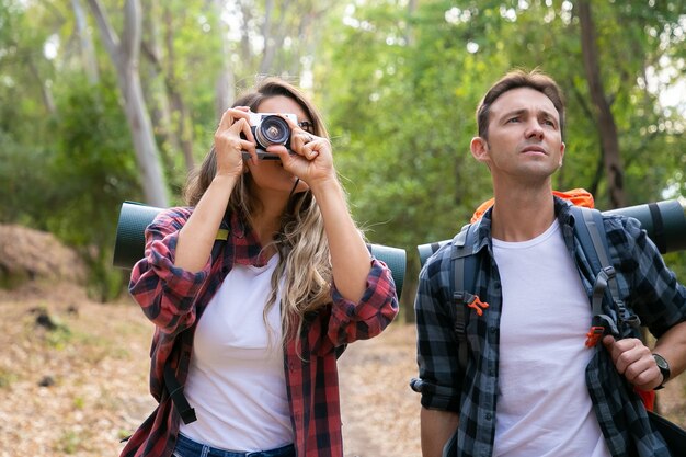 Caucasian young couple hiking in forest and taking photo with camera. Thoughtful male traveler standing near woman and looking at landscape. Backpacking tourism, adventure and summer vacation concept