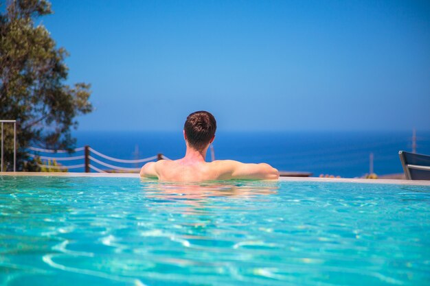 Caucasian young man in the infinity pool watching the ocean view, relaxing, and enjoying his life