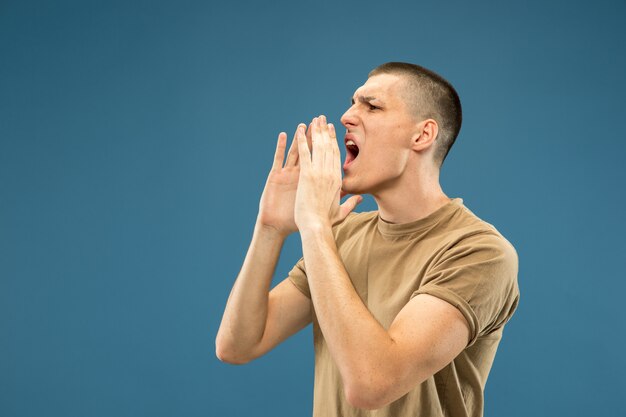 Caucasian young man's half-length portrait on blue studio background. Beautiful male model in shirt. Concept of human emotions, facial expression, sales, ad. Calling somebody, screaming.