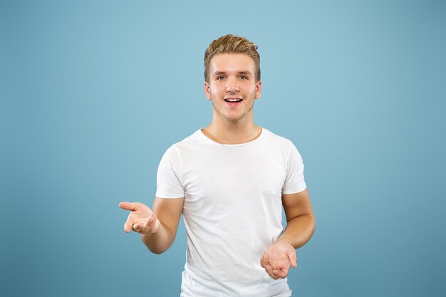 Caucasian young man's half-length portrait on blue studio background. Beautiful male model in shirt. Concept of human emotions, facial expression, sales, ad. Pointing and showing something.