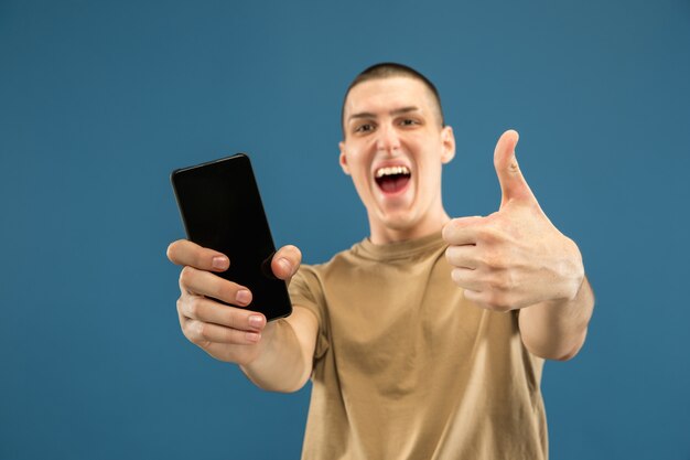 Caucasian young man's half-length portrait on blue studio background. Beautiful male model in shirt. Concept of human emotions, facial expression, sales, ad. Showing phone screen, looks astonished.