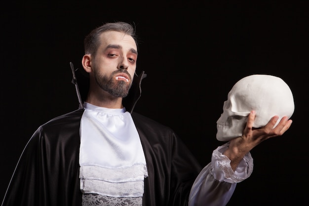 Free Photo caucasian young man with scary face and dracula costume looking into the camera and holding a skull. scary and dangerous man with vampire eyes.