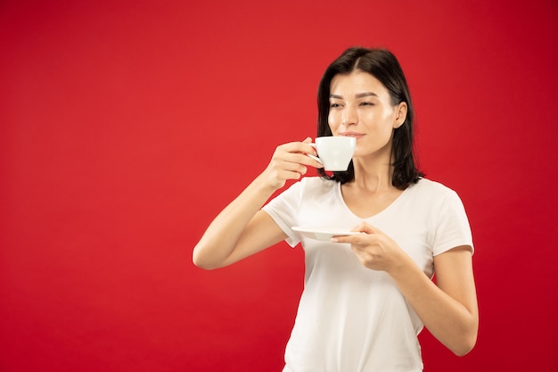 Caucasian young woman's half-length portrait on red studio background. Beautiful female model in white shirt. Concept of human emotions, facial expression. Enjoys drinking coffee or tea, looks calm.