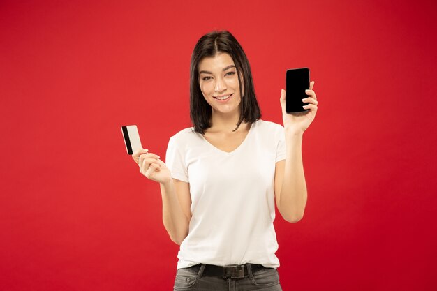 Caucasian young woman's half-length portrait on red studio background. Beautiful female model in white shirt. Concept of human emotions, facial expression, sales. Online payment bill or purchases.