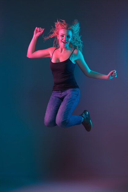 Caucasian young woman's portrait on gradient background in neon light. Beautiful female model with unusual look. Concept of human emotions, facial expression, sales, ad. Jumping, smiling.