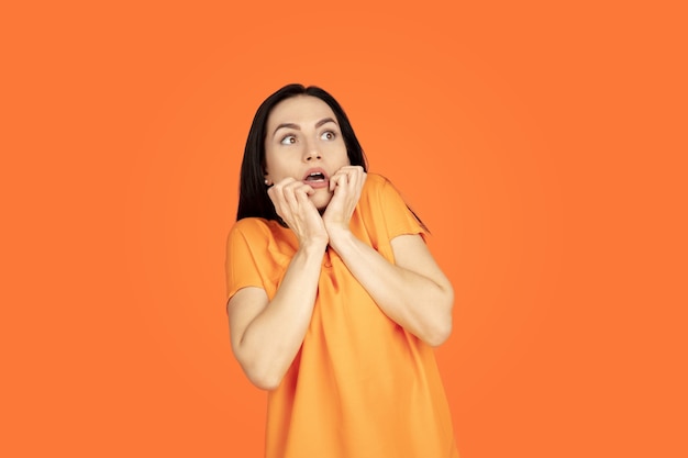 Caucasian young woman's portrait on orange studio background. Beautiful female brunette model in shirt. Concept of human emotions, facial expression, sales, ad. Copyspace. Looks scared.