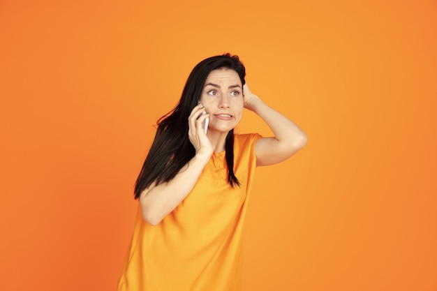 Caucasian young woman's portrait on orange studio background. Beautiful female brunette model in shirt. Concept of human emotions, facial expression, sales, ad. Copyspace. Talking on phone.