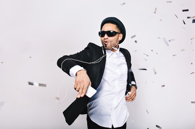 Free photo celebrating brightful great party of excited handsome guy in suit, hat, black sunglasses having fun in tinsels. fashionable look, listening to music, dancing, singing, relaxing.