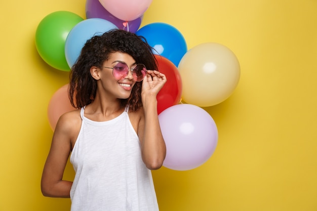 Celebration Concept - Close up Portrait happy young beautiful african woman in black t-shirt smiling with colorful party balloon. Yellow Pastel studio Background.