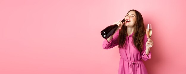 Celebration and holidays concept Happy carefree woman drinking champagne from bottle with pleased smile relaxing on party holding glass standing over pink background