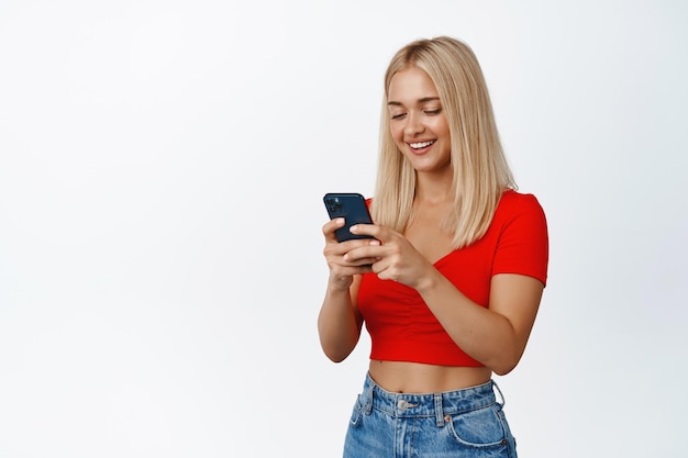 Cellular technology Young blond woman using mobile phone and smiling messaging or placing an oder on smartphone white background