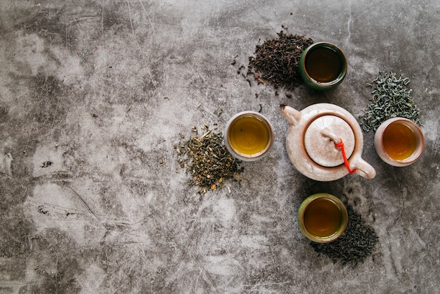 Free photo ceramic teapot surrounded with dried herbs and teacups on concrete backdrop