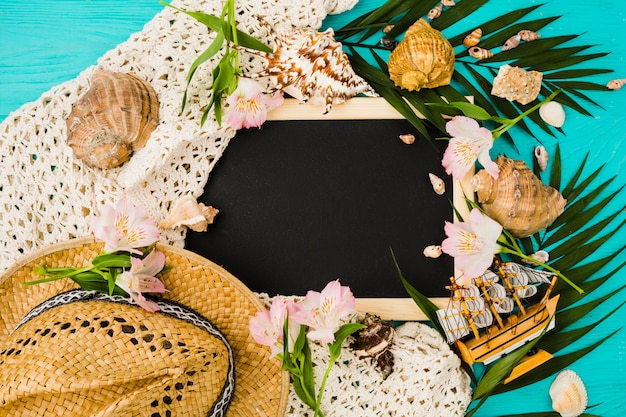 Chalkboard between plant leaves with flowers near seashells and hat