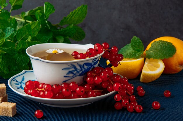 chamomile tea in cup and sauce with lemon with slices, fresh red berries, brown sugar and leaves on grey stucco and dark blue placemat background. vertical side view