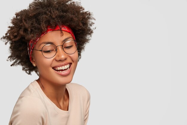 Charismatic happy black curly young lady looks down with broad smile, wears transparent glasses, red headband, laughs at something funny, poses against white wall, blank copy space on right side.