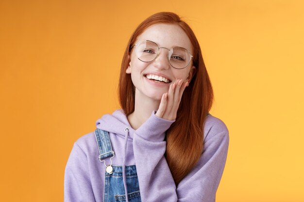 Charismatic happy tender redhead smiling european girl 20s glasses hoodie denim overalls having fun enjoying summer holidays chilly evening laughing joyfully touch pure clear skin, orange background.