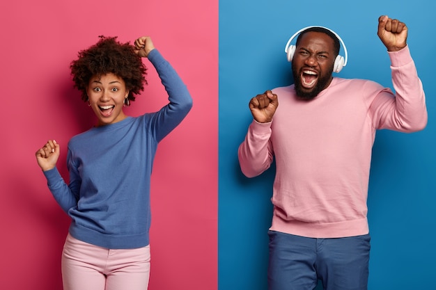 Charismatic joyful Afro American woman and man pull hands up and dance happily with rhythm of music, wear headphones, pose against blue and pink space. People