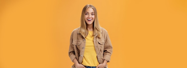 Free photo charismatic tanned woman in corduroy jacket and yellow tshirt ready for chilly autumn walk with friends smiling joyfully gazing entertained at camera holding hand in pockets casually over orange wall