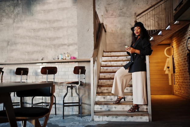 Free photo charming african american woman model in black jacket hat and waist bag relaxing in cafe during free time with mobile phone