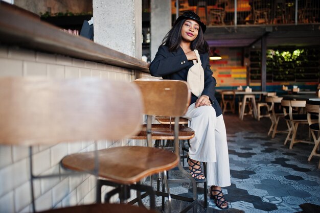 Charming african american woman model in black jacket hat and waist bag relaxing in cafe during free time