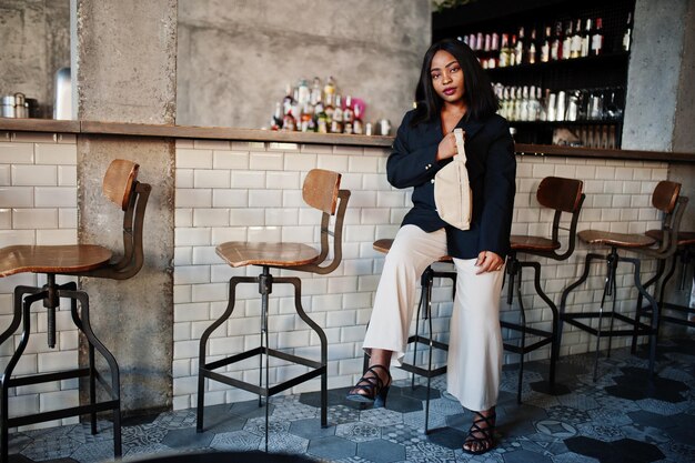 Charming african american woman model in black jacket and waist bag relaxing in cafe during free time