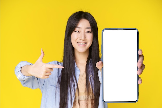 Charming asian girl with smartphone in hand pointing finger on white screen Great offer Mobile app advertisement product placement