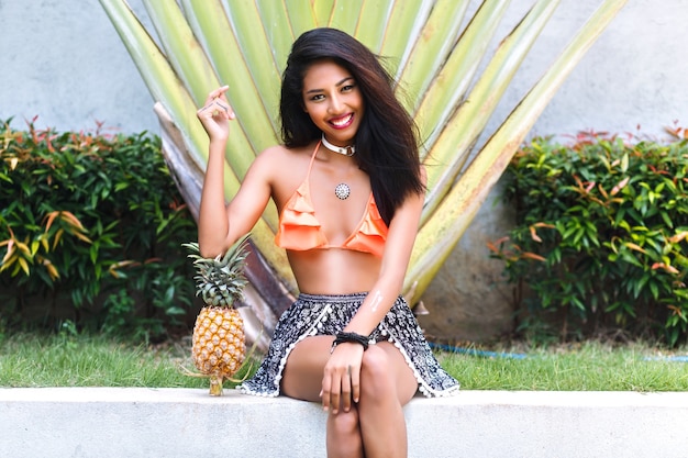 Charming asian girl with sparkle fake tattoo on hand sitting in lotus pose and holding pineapple. Outdoor portrait of relaxed brunette woman doing yoga in exotic park with palm trees.