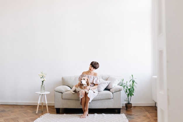 Charming barefooted girl sitting on gray sofa between little table and green plant, looking at beagle dog on her knees