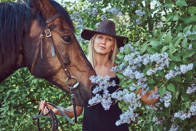 Free Photo charming beautiful blonde wearing black clothes and hat standing with a horse in the flower garden.