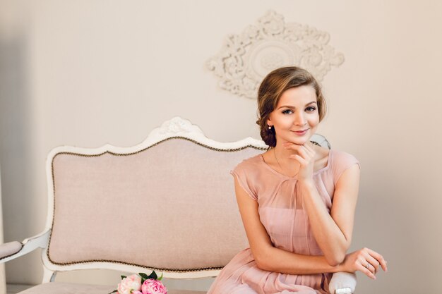 Charming blond girl sitting on a sofa. She has braided hair and holds her hand near chin.