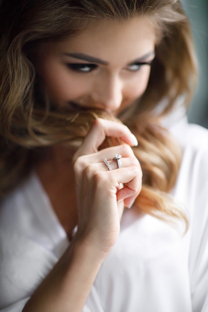 Free photo charming blonde bride with curls and shiny skin poses in white silk robe in the room