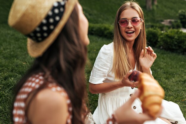 Charming blonde long-haired girl in red sunglasses and white dress holds apple, sits on rug on grass and talks to her brunette curly friend