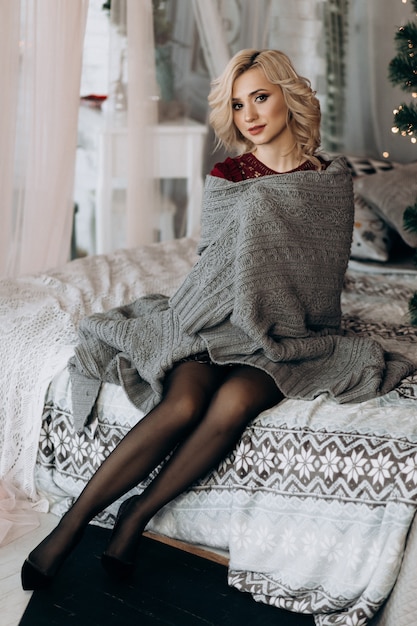 Free photo charming blonde woman envelopes herself in grey plaid sitting on a bed before a christmas tree