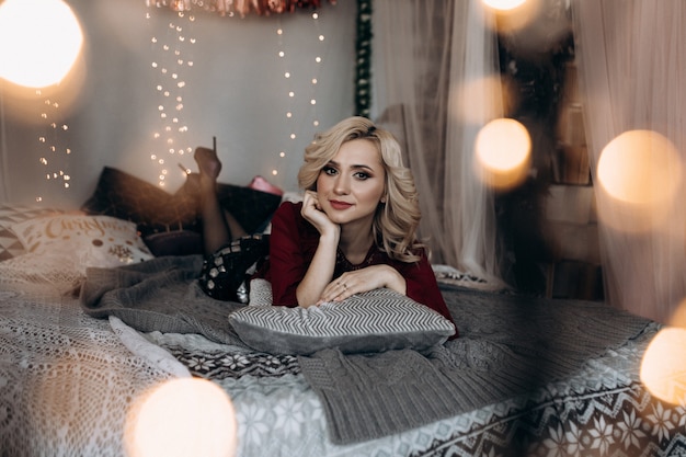 Charming blonde woman envelopes herself in grey plaid sitting on a bed before a Christmas tree